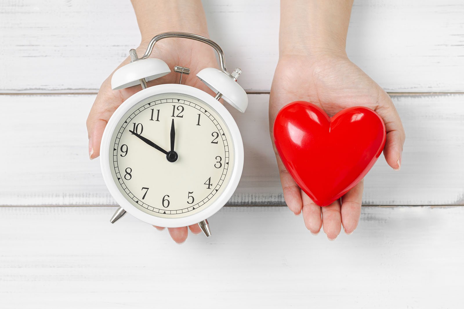 Woman Hands Hold Alarm Clock and Red Heart on white wood background